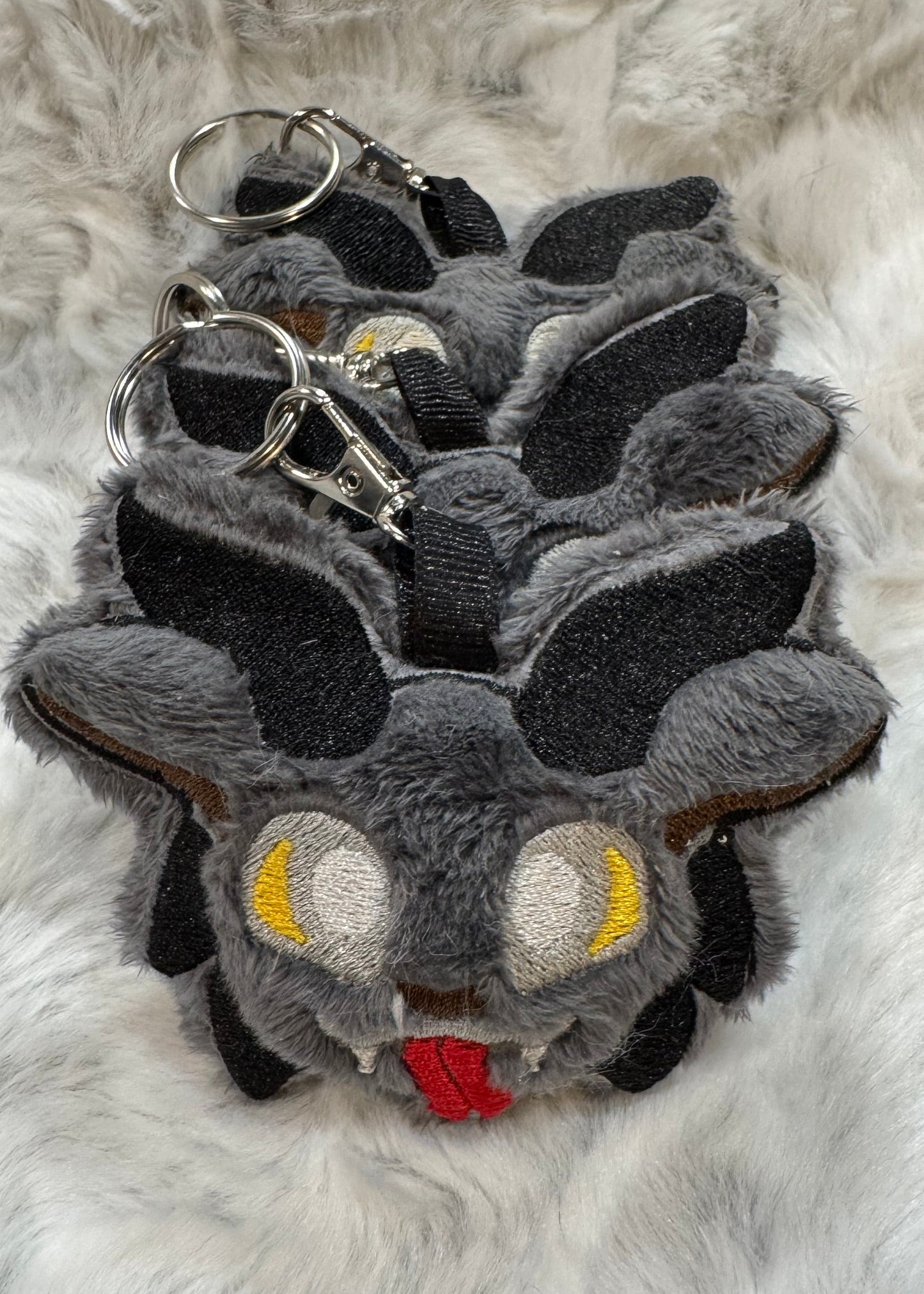 A group of plush Krampus keychains with gray faux fur, black horns, yellow embroidered eyes, and red tongues, arranged in a row on soft white fabric.