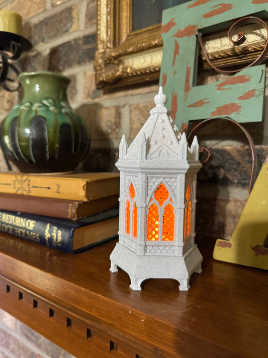 White stone gothic cathedral lantern with the orange lattice window glowing, spire finial top, on a wood mantel beside antique books.
