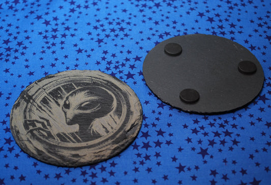 Round slate coaster with an alien peering out of a portal and the bottom also shown, with rubber protective feet.