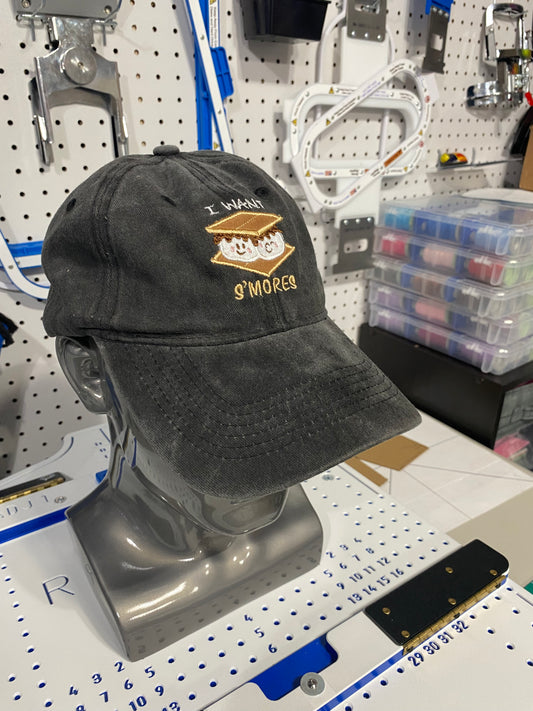 A black washed baseball cap featuring a gold embroidered s'mores graphic with the text 'I WANT S'MORES' displayed on a gray mannequin head. The background shows a workshop or studio setting with a pegboard, tools, and storage bins.