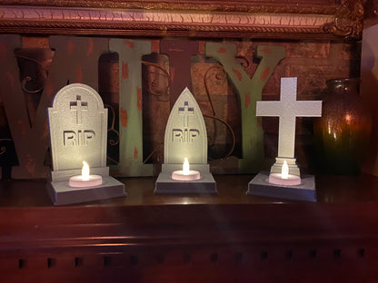 Set of three 3D printed tombstone tea light holders on a mantel: rounded tombstone, gothic arch, and cross, each lit by an LED tea light