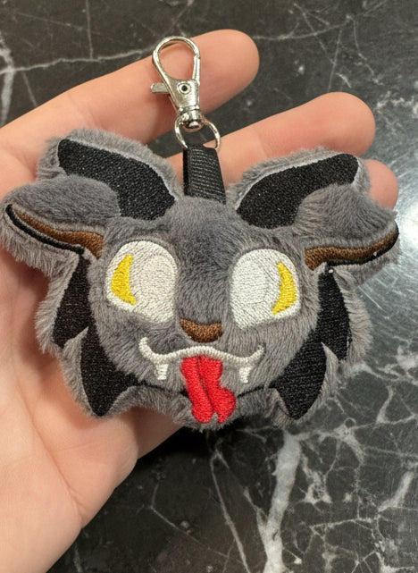 Plush Krampus keychain with gray faux fur, embroidered yellow eyes, red tongue, and black horns, attached to a silver clasp, displayed on rustic wooden background.
