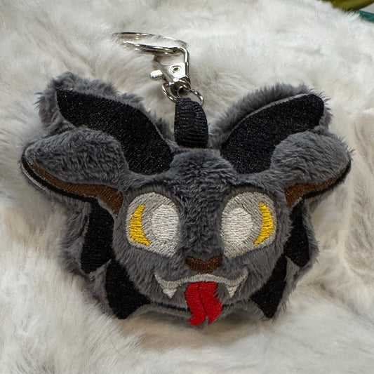 lose-up of a plush Krampus keychain with gray faux fur, yellow embroidered eyes, red tongue, and black horns, attached to a silver clasp, resting on white faux fur.
