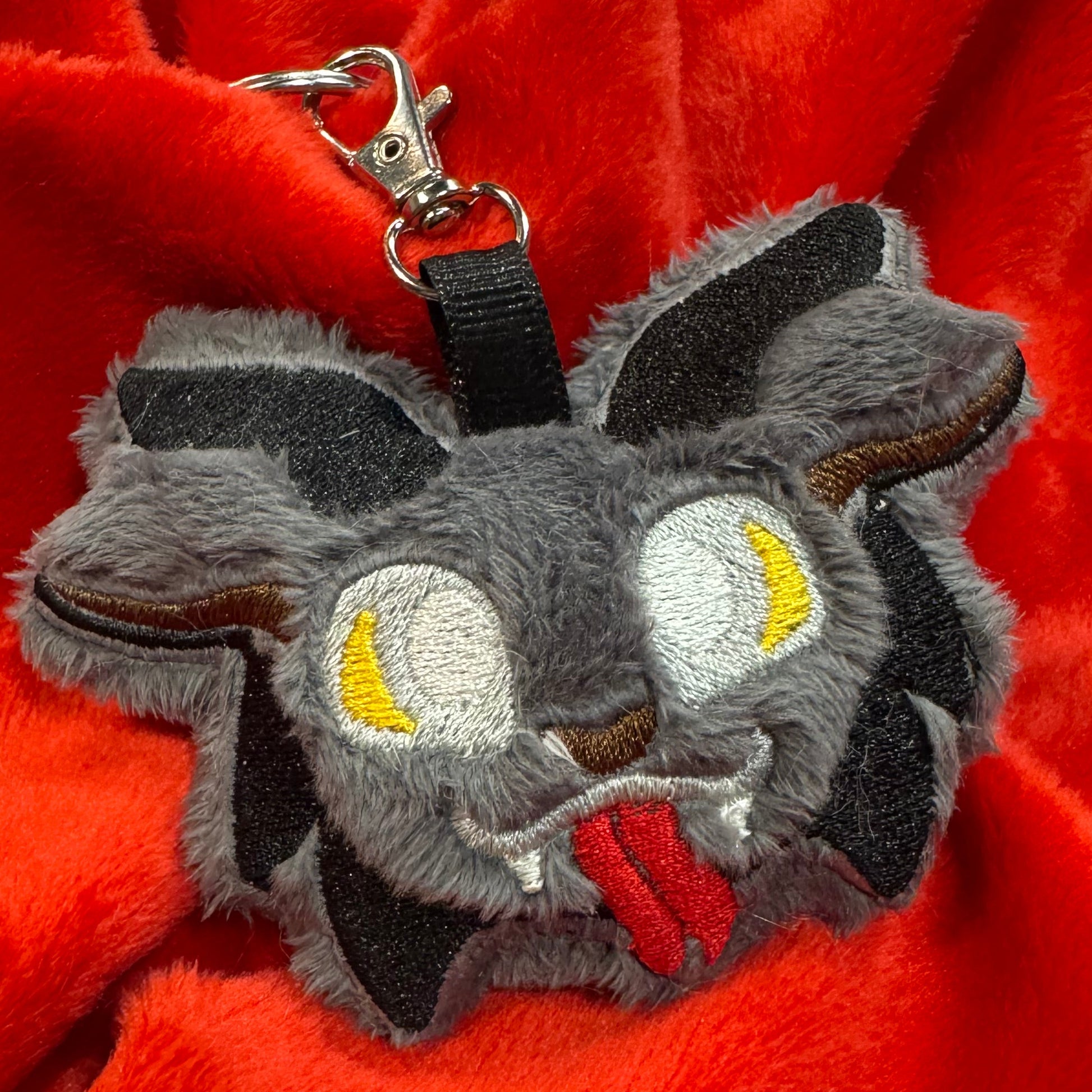 Close-up of a plush Krampus keychain featuring fuzzy gray faux fur, embroidered yellow eyes, red tongue, and small black horns, attached to a silver key clasp, resting on bright red fabric.