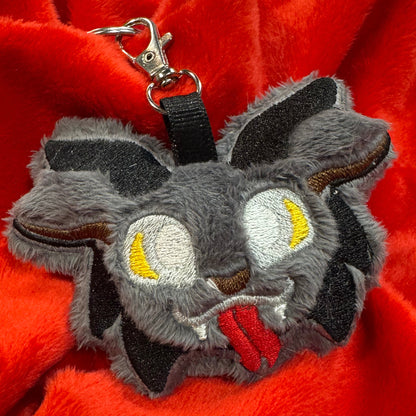 Close-up of a plush Krampus keychain featuring fuzzy gray faux fur, embroidered yellow eyes, red tongue, and small black horns, attached to a silver key clasp, resting on bright red fabric.
