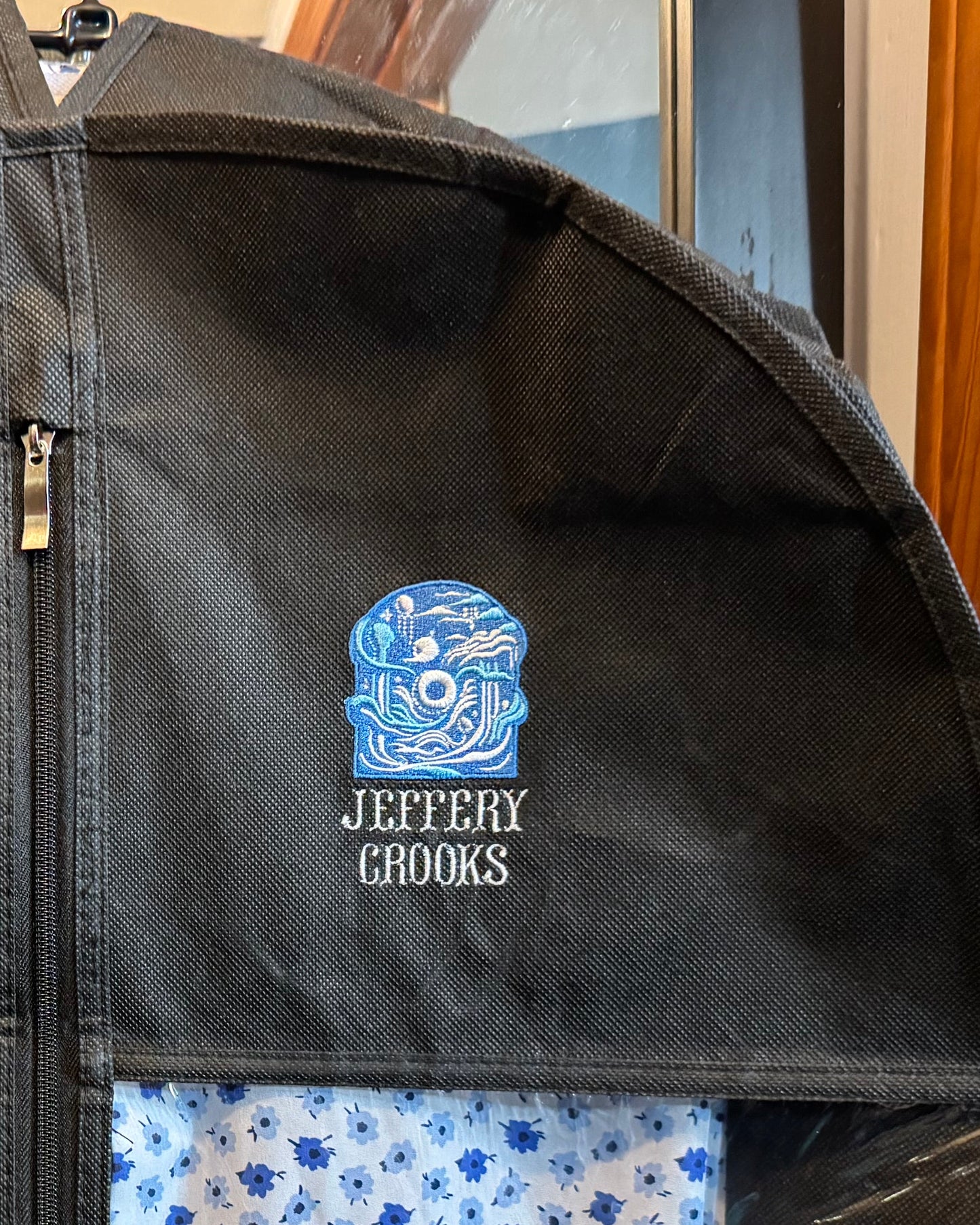 A black zippered garment storage bag hanging indoors, with a blue and white patterned fabric insert visible through a front window and the ItsNotNought logo on the upper right side with name "Jeffery Crooks" underneath the logo.