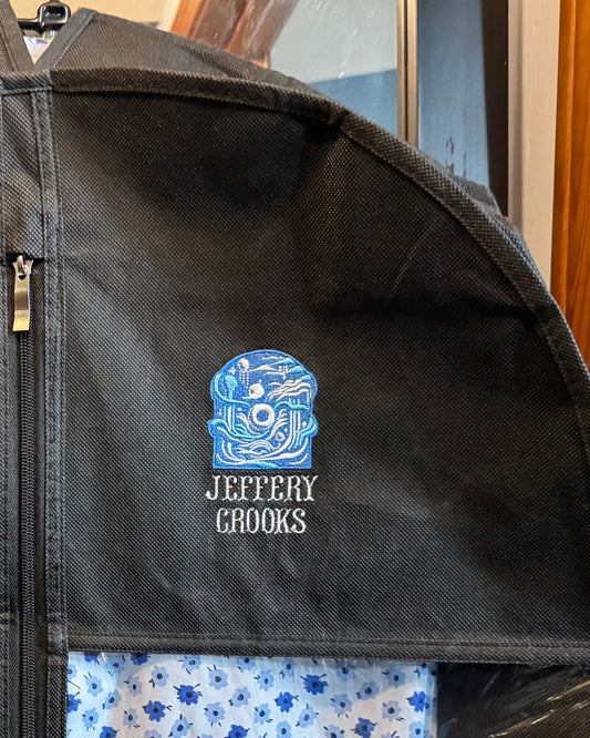 A black zippered garment storage bag hanging indoors, with a blue and white patterned fabric insert visible through a front window and the ItsNotNought logo on the upper right side with name "Jeffery Crooks" underneath the logo.