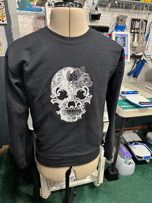 A black long-sleeve crewneck sweatshirt displayed on a dress form mannequin in a workshop or studio setting. The sweatshirt features a large white and silver graphic of a decorative sugar skull on the front.