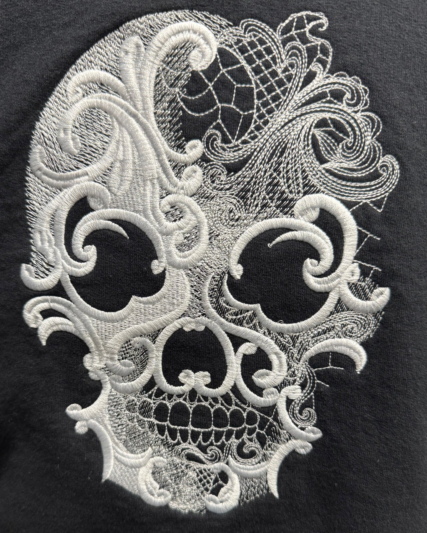 A close-up of a black garment featuring a large white embroidered lace-patterned skull design on the front.