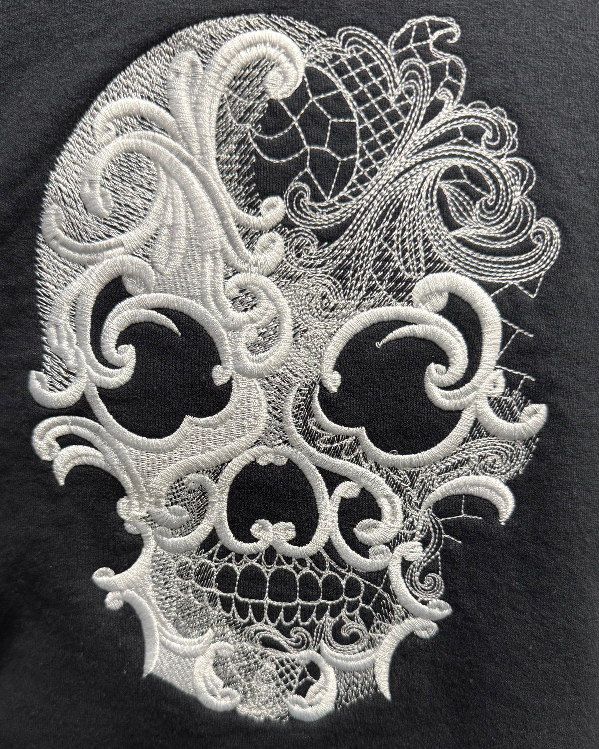 A close-up of a black garment featuring a large white embroidered lace-patterned skull design on the front.