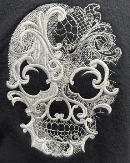 A close-up of a black garment featuring a large white embroidered lace-patterned skull design on the front.