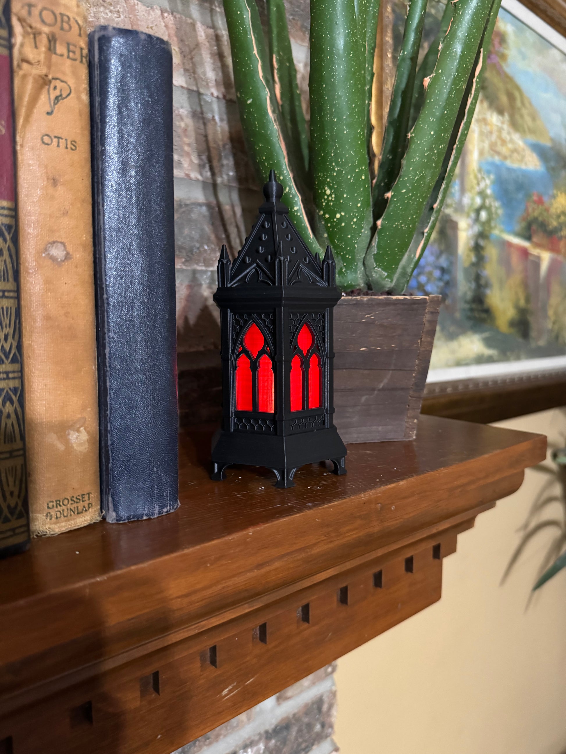 Black gothic cathedral lantern with red glowing arched windows and a spire finial top, on a wood mantel next to antique books.