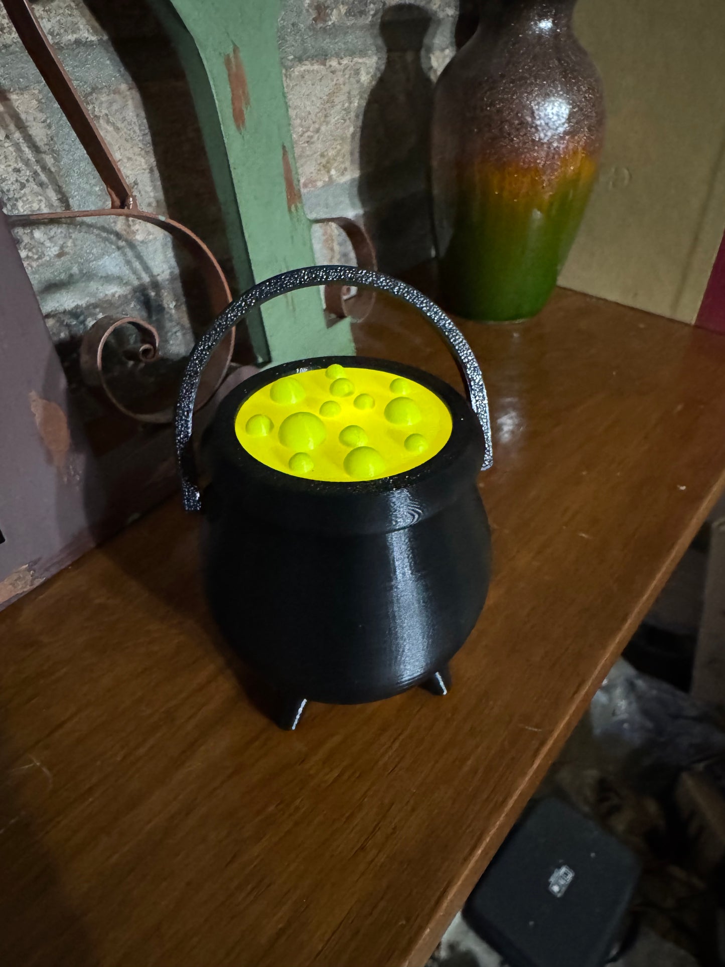 Mini black cauldron with a glowing green potion top and raised handle, 3D printed Halloween decor on a wood shelf