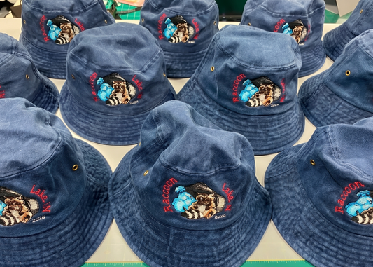 Multiple blue bucket hats with embroidered raccoon designs and red text, arranged on a white table.