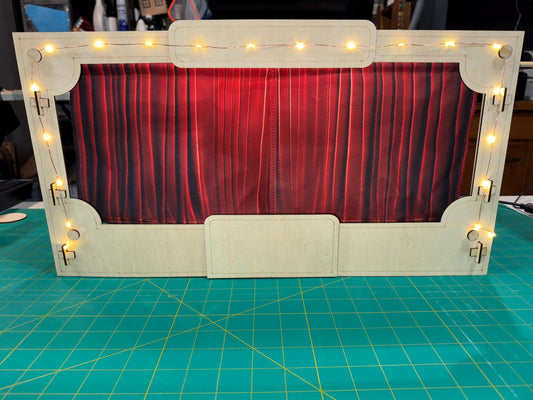 Front view of the assembled tabletop puppet theater with closed red curtains and warm fairy lights along the wooden frame